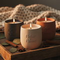 Three lit scented candles in matte ceramic vessels on a rustic wooden tray with eucalyptus leaves and warm golden lighting